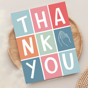 A colorful "thank you" card with a grid design featuring pastel blocks, each containing one letter with prayer hands, placed on a wooden surface.