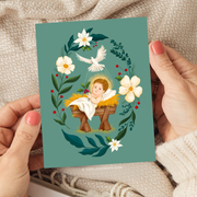 Christmas card with Baby Jesus in a manger surrounded by florals and holly, holding a sprig of holly in His hand.