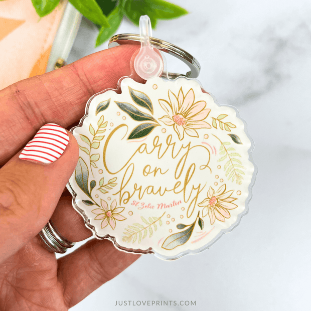 Keychain with floral design and 'Carry on bravely' text held by a hand.
