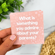 Hand holding a pink card with text 'What is something you admire about your parents?' on a light background.