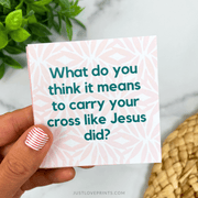 Hand holding a card with a question about carrying one's cross like Jesus did, with a plant and woven basket in the background.