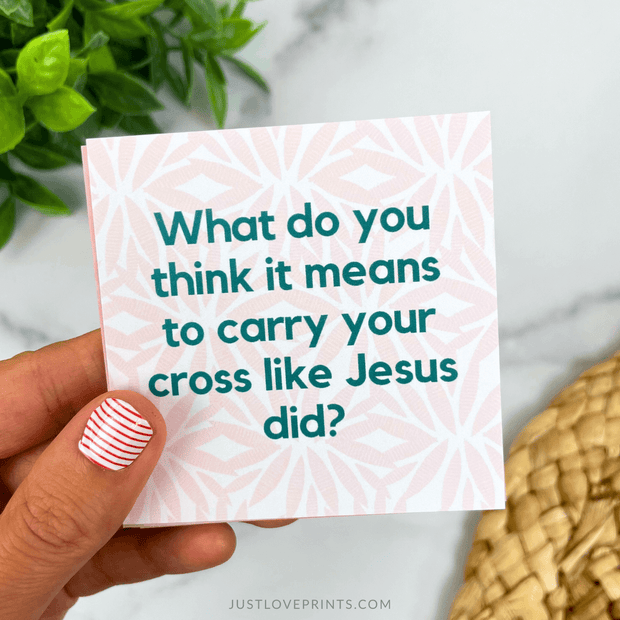 Hand holding a card with a question about carrying one's cross like Jesus did, with a plant and woven basket in the background.
