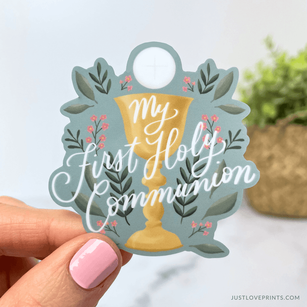 The image features a decorative sticker with the text "My First Holy Communion" written in elegant script. The design includes a golden chalice with a white Eucharistic host above it, adorned with a faint cross. Surrounding the chalice are green leaves and small pink flowers, giving it a delicate and celebratory feel. The background is a soft muted blue, making the text and imagery stand out. This type of sticker is commonly used for First Communion celebrations, scrapbooking, or as a keepsake. 