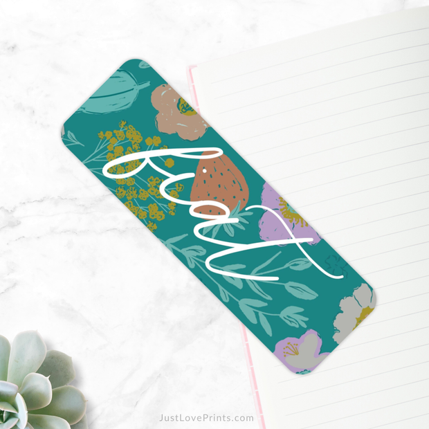 A teal-colored bookmark featuring a vibrant floral pattern with various flowers and leaves in shades of pink, yellow, purple, and green is placed diagonally on a white marble surface. The word "fiat" is written in a large, white, cursive font across the bookmark. 