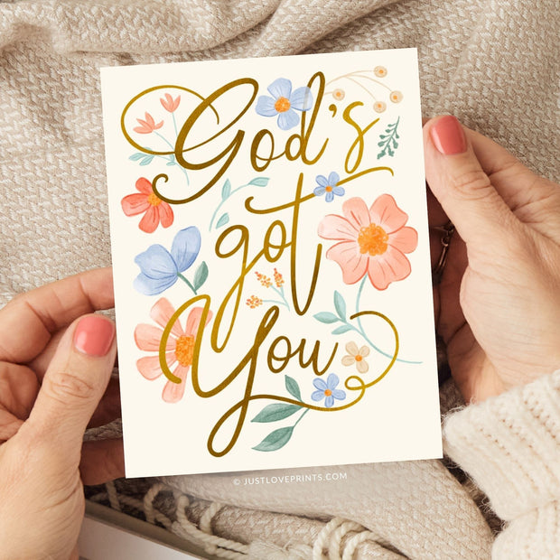 This card design radiates warmth and reassurance with the message "God's got you" in a bold, flowing gold script. Surrounding the text are soft, hand-painted floral elements in hues of peach, blue, and cream, complemented by green leaves. The arrangement of flowers and gentle colors creates an uplifting and peaceful aesthetic, perfect for conveying comfort and encouragement with a faith-centered focus.