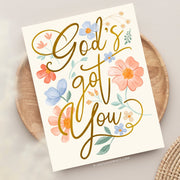 This card design radiates warmth and reassurance with the message "God's got you" in a bold, flowing gold script. Surrounding the text are soft, hand-painted floral elements in hues of peach, blue, and cream, complemented by green leaves. The arrangement of flowers and gentle colors creates an uplifting and peaceful aesthetic, perfect for conveying comfort and encouragement with a faith-centered focus.