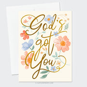 This card design radiates warmth and reassurance with the message "God's got you" in a bold, flowing gold script. Surrounding the text are soft, hand-painted floral elements in hues of peach, blue, and cream, complemented by green leaves. The arrangement of flowers and gentle colors creates an uplifting and peaceful aesthetic, perfect for conveying comfort and encouragement with a faith-centered focus.