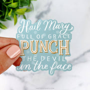 This playful sticker reads, "Hail Mary, full of grace, punch the devil in the face," set against a soft blue background with bold and elegant typography.