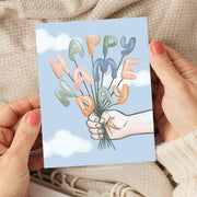 This card design celebrates a "Happy Name Day" with playful charm. It features a hand holding a bundle of balloon letters that spell out the greeting in cheerful pastel colors, including soft oranges, greens, and blues. The background is a light sky blue with soft white clouds, giving a whimsical, uplifting feel to the design. Perfect for marking a special Name Day with a joyful, personalized touch.