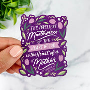 This sticker displays St. Thérèse of Lisieux's quote, "The loveliest masterpiece of the heart of God is the heart of a mother," surrounded by floral accents on a rich purple background.