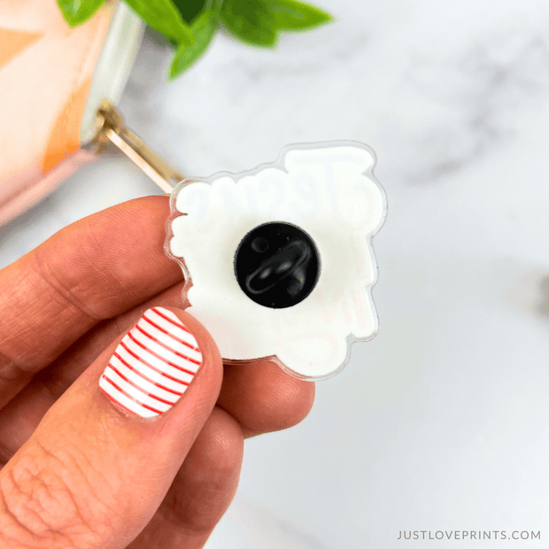 White pin with black pin backing held by a hand against a light background.