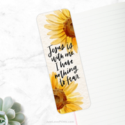 A white bookmark featuring two large yellow sunflowers is placed diagonally on a white marble surface. The text on the bookmark reads, "Jesus is with me. I have nothing to fear." The bottom left corner of the image shows a small green succulent, and in the background, there is an open lined notebook with a pink edge. The website "JustLovePrints.com" is printed at the bottom of the image.