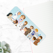 A bookmark with a light blue background is placed diagonally on a white marble surface. The bookmark features cartoon-style illustrations of various saints and religious figures, including Mother Teresa, among others, all standing in a row. 