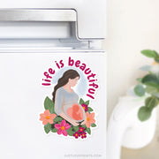 Car magnet with illustration of a pregnant mother surrounded by flowers and the words “Life is Beautiful.”