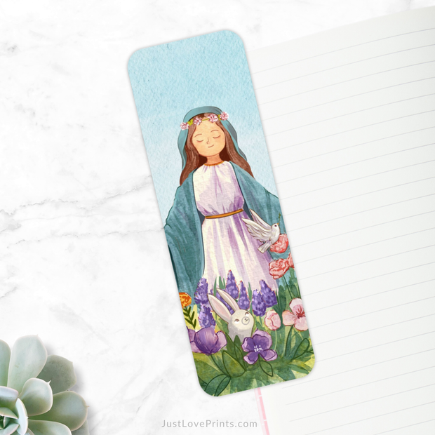 A bookmark with a light blue background is placed diagonally on a white marble surface. The bookmark features an illustration of Mother Mary, depicted with a serene expression, wearing a white dress with a light blue cloak and a floral crown. She is standing among colorful flowers with a white dove on her right and a small gray bunny on her left. 