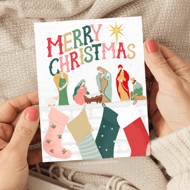 This festive card design spreads holiday cheer with a vibrant "Merry Christmas" message at the top in bold, colorful letters. Below, a classic nativity scene features Mary, Joseph, Baby Jesus in the manger, an angel, shepherds, and the Magi, all illustrated in warm, earthy tones. Above a cozy fireplace, stockings in various colors and patterns are hung with care, adding a touch of traditional Christmas spirit. The scene is topped with a bright star, symbolizing the Star of Bethlehem. 