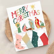 This festive card design spreads holiday cheer with a vibrant "Merry Christmas" message at the top in bold, colorful letters. Below, a classic nativity scene features Mary, Joseph, Baby Jesus in the manger, an angel, shepherds, and the Magi, all illustrated in warm, earthy tones. Above a cozy fireplace, stockings in various colors and patterns are hung with care, adding a touch of traditional Christmas spirit. The scene is topped with a bright star, symbolizing the Star of Bethlehem.