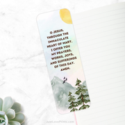 A white bookmark with an illustration of a mountain landscape, featuring green trees and birds flying in the sky, is placed diagonally on a white marble surface. The text on the bookmark reads, "O JESUS, THROUGH THE IMMACULATE HEART OF MARY, I OFFER YOU MY PRAYERS, WORKS, JOYS, AND SUFFERINGS OF THIS DAY. AMEN." The top right corner shows a bright yellow sun. 