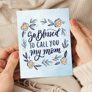 A greeting card with elegant script reading "so blessed to call you my mom" surrounded by a floral design with yellow and pink flowers and navy leaves,