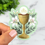 Sticker with golden chalice, Eucharist host with IHS letters, and white lilies around it.