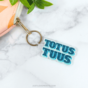 Keychain with 'TOTUS TUUS' text on a white surface with a plant in the corner.