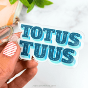Hand holding a clear keychain with blue text 'TOTUS TUUS' against a white background.