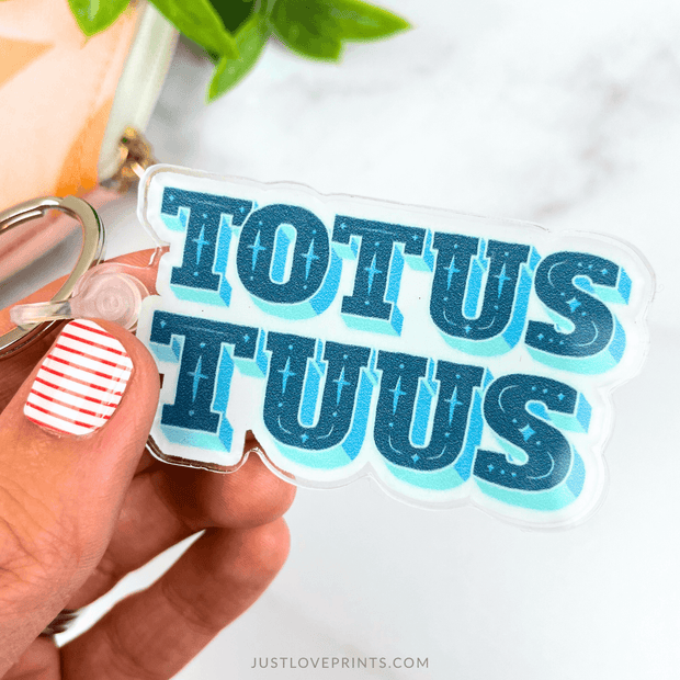 Hand holding a clear keychain with blue text 'TOTUS TUUS' against a white background.