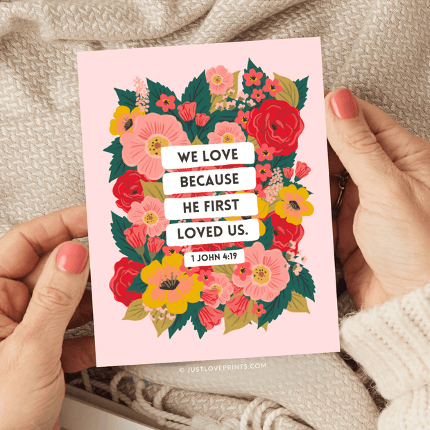 Floral pink greeting card featuring the Bible verse:
"We love because He first loved us." – 1 John 4:19.