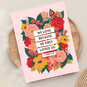 Floral pink greeting card featuring the Bible verse:
"We love because He first loved us." – 1 John 4:19.