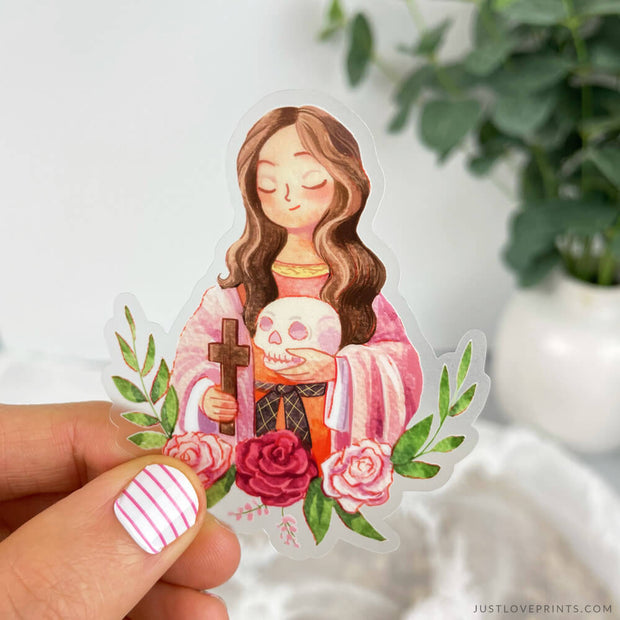 A watercolor sticker image of St. Mary Magdalene holding a cross and skull with a laurel wreath and roses beneath her.  