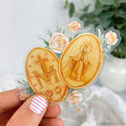 this gold miraculous medal sticker shows both sides of a miraculous medal with roses in the background. the medal is in gold. 